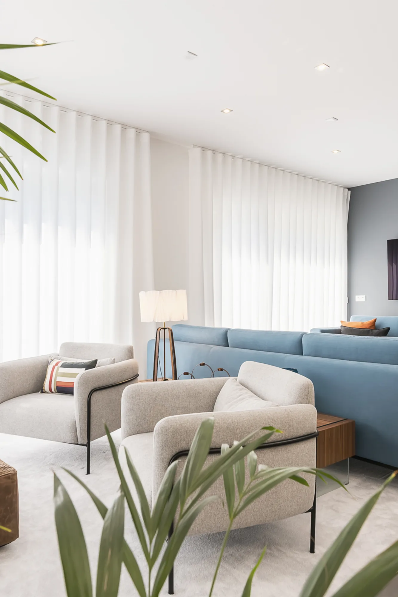 Sala de estar com sofá azul, cadeirões claros e cortinados translúcidos a toda a altura.