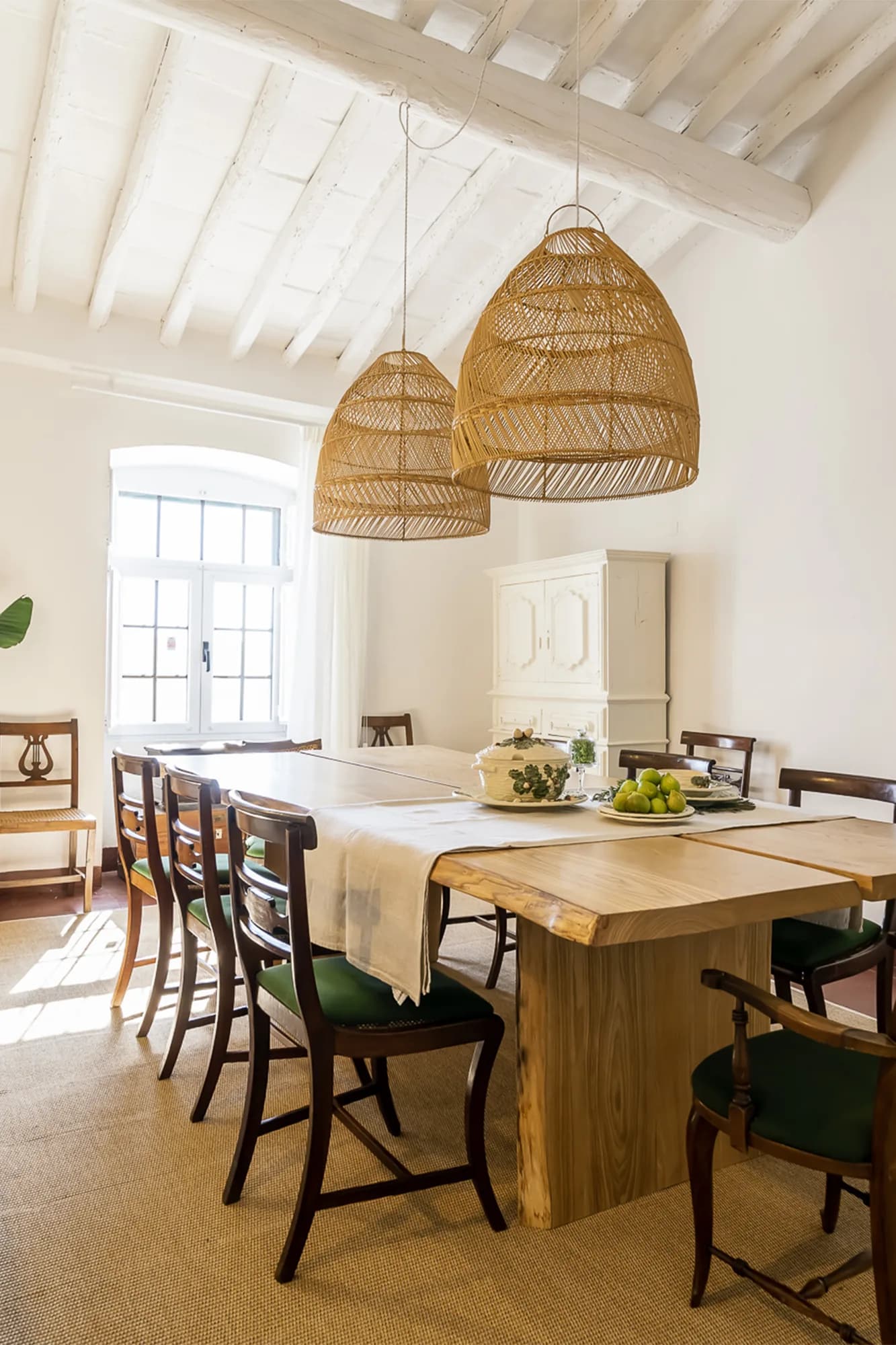 Sala de jantar sob tecto de vigas caiadas, com mesa em madeira maciça, cadeiras antigas e suspensões em fibra natural.